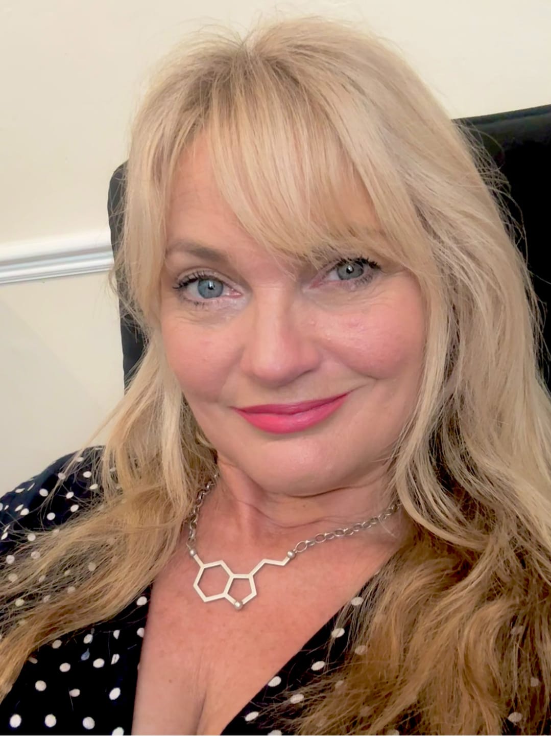 Woman with blonde hair, smiling, wearing a molecular structure necklace and polka dot top.